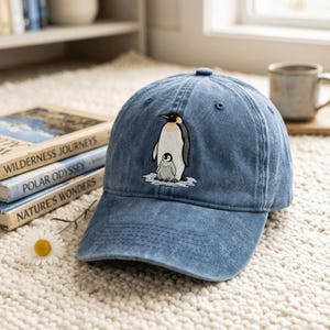 May include: A blue denim baseball cap featuring an embroidered graphic of an adult emperor penguin standing with a small chick on a patch of white ice. The cap has a structured crown, a curved brim, and a button at the top centre of the panels.