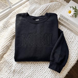 以下が含まれることがあります： 胸元に同色の黒いカレッジフォントでMIMIという文字が刺繍された、黒いクルーネックのスウェットシャツ。左袖口にはSophiaという名前が黒い筆記体で刺繍されており、デザインを完成させています。