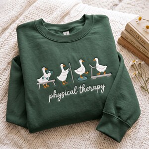 May include: A dark green crewneck sweatshirt featuring an embroidered graphic of four white geese performing physical therapy exercises, including lifting dumbbells, using resistance bands, balancing on a disc, and walking on a treadmill.