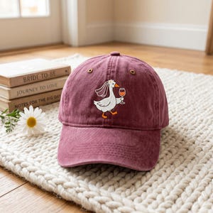 May include: A maroon baseball cap featuring an embroidered graphic of a white goose wearing a bridal veil and holding a glass of orange wine. The cap has a curved brim, a button on top, and metal eyelets for ventilation on the crown panels.