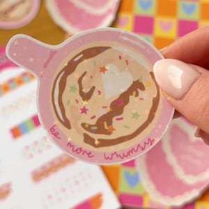 Puede incluir: Una pegatina rosa con un diseño de taza de café, con goteo de chocolate, chispas y un corazón. La pegatina tiene las palabras "be more whimsy" y la sostiene una persona. Otras pegatinas con diseños similares están en el fondo.