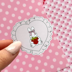 Peut inclure: Un autocollant en forme de cœur représentant un lapin blanc tenant une fraise rouge. Le lapin a un nœud rose et est centré sur un cœur rose clair avec un bord à volants. Le fond est rose avec des pois blancs et un motif de cœurs rouges et blancs.