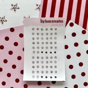 May include: A sheet of alphabet stickers with the text "by han creates" and "Alphabet Brede". The stickers are small, round, and feature letters A through Z. The background includes pink and white patterned paper with stars, stripes, and polka dots.