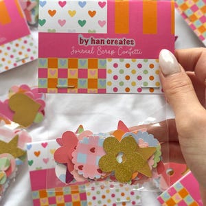 May include: A clear bag containing assorted paper confetti in flower and star shapes. The confetti is packaged with a pink card featuring the text "by han creates Journal Scrap Confetti". The card has patterned sections.