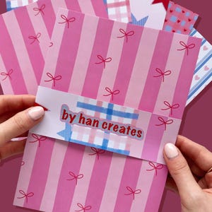 May include: A stack of pink striped cards with red bow accents and the text "by han creates" on a decorative band. Additional cards feature patterns of hearts, stars, and gingham. The cards are held by a person.