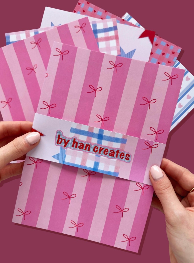 May include: A stack of pink striped cards with red bow accents and the text "by han creates" on a decorative band. Additional cards feature patterns of hearts, stars, and gingham. The cards are held by a person.