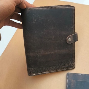 May include: Dark brown leather journal with a snap closure. The journal has a textured surface and visible stitching along the edges. A smaller, blue leather item is visible in the bottom right corner.