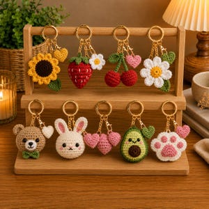 May include: A collection of nine handmade crochet keychains hanging on a wooden display stand. The designs include a sunflower, strawberry, cherries, daisy, bear, rabbit, heart cluster, avocado, and a paw print, each with a gold metal ring.