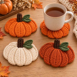 Può includere: Un set di tre sottobicchieri all'uncinetto a forma di zucca, due arancioni e uno color crema, ciascuno con un gambo marrone scuro e una piccola foglia verde. Un sottobicchiere arancione sostiene una tazza in ceramica maculata.