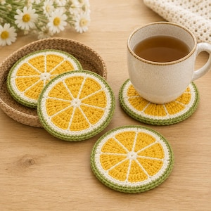 May include: Four round crochet coasters designed to look like citrus slices. Each coaster features a bright yellow center with white segments and a green outer border. One coaster sits under a speckled ceramic mug filled with tea.