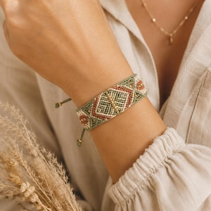 May include: A woven macrame bracelet featuring a geometric pattern in sage green, terracotta, and cream thread. The design includes a central vertical line of small gold-toned metallic beads integrated into the intricate knotwork pattern.