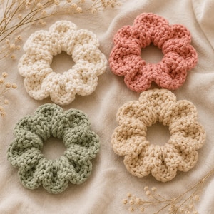 May include: Four circular hair scrunchies made of thick, crocheted yarn. The set includes one white, one dusty pink, one sage green, and one beige scrunchie. Each item features a scalloped, flower-like edge design with a soft, textured finish.