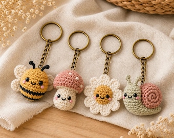 May include: Four crochet keychains featuring a yellow and black bee, a pink and white mushroom, a white and yellow daisy, and a light green snail with a pink shell. Each item is attached to a bronze-colored metal ring and a short chain link.