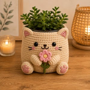 May include: A beige crochet plant pot cover shaped like a cat, featuring pink inner ears, pink paw pads, and a small pink flower held by two front paws. The cat face has black embroidered eyes, a nose, and whiskers, holding a green stem.
