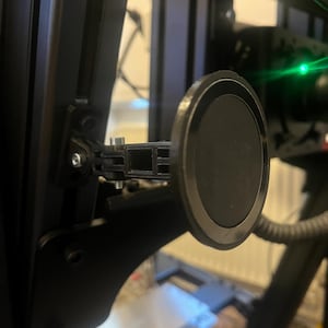Könnte beinhalten: Ein schwarzer, verstellbarer Montagebügel mit einer kreisförmigen, schwarzen Magnetoberfläche. Der Bügel ist an einem schwarzen Metallrahmen befestigt, wahrscheinlich für ein Gaming- oder Sim-Racing-Setup. Das Design wirkt funktional und industriell.