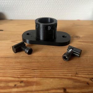 Puede incluir: Componentes de hardware negros impresos en 3D, que incluyen una pieza cilíndrica con agujeros, una base rectangular con orificios de montaje y dos pernos en T. Los artículos están dispuestos sobre una superficie de madera.