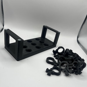 Pode incluir: Organizador preto impresso em 3D com vários orifícios circulares e uma pilha separada de pequenos clipes pretos. O organizador tem uma forma retangular com suportes verticais. Os clipes têm um design único.