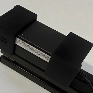 May include: A black electronic device with a barcode label and a logo. The device has a rectangular shape with a series of black brackets and a black cable. The barcode label contains the numbers 104.022.00173-234102741.