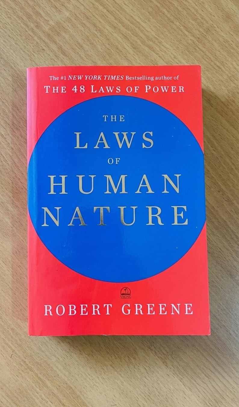 May include: A hardcover book titled The Laws of Human Nature by Robert Greene. The cover features a bright red background with a large blue circle in the center. The text is printed in white and gold, including the author name and book title.