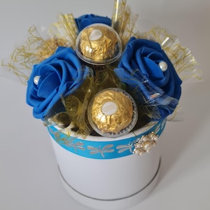 May include: A white round gift box with a blue ribbon featuring dragonfly designs. The box holds blue fabric roses and gold-wrapped chocolate candies. Decorative gold and clear cellophane and a pearl brooch complete the arrangement.