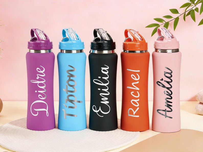 Puede incluir: Cinco botellas de agua personalizadas en morado, azul, negro, naranja y rosa. Cada botella tiene un nombre plateado impreso verticalmente. Las botellas tienen una tapa abatible y una base de acero inoxidable. Los nombres son Deidre, Tipton, Emilia, Rachel y Amelia.