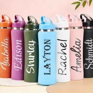 Può includere: Una collezione di borracce personalizzate in vari colori, tra cui arancione, viola, verde, blu, bianco, rosa e nero. Ogni borraccia presenta un nome in un elegante carattere corsivo. Le borracce hanno un coperchio ribaltabile e sono progettate per l'idratazione.