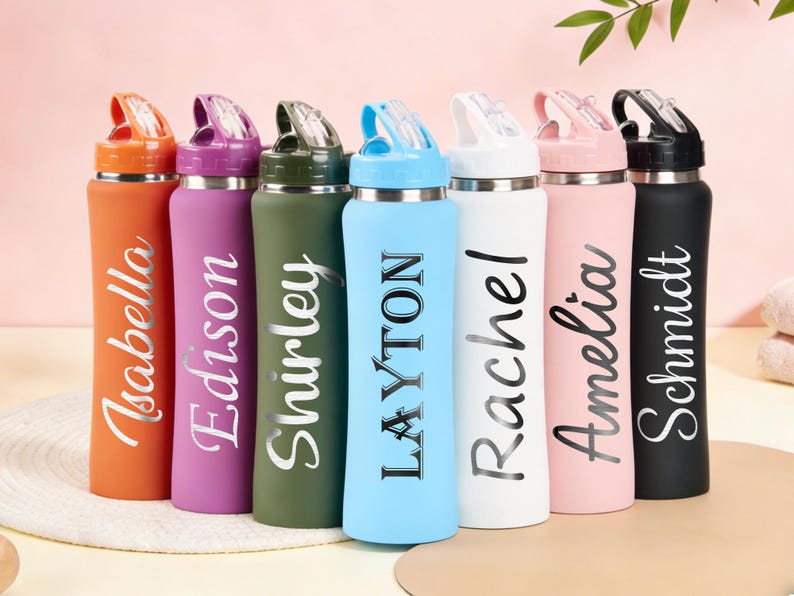 Puede incluir: Una colecci&oacute;n de botellas de agua personalizadas en varios colores, incluyendo naranja, morado, verde, azul, blanco, rosa y negro. Cada botella presenta un nombre en una fuente elegante. Las botellas tienen una tapa abatible y est&aacute;n dise&ntilde;adas para la hidrataci&oacute;n.