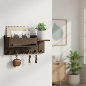Könnte beinhalten: Ein dunkelbrauner Wand-Schlüsselhalter aus Holz mit einem Regal, einem Postschlitz und sechs Haken. Auf dem Regal befinden sich eine kleine Topfpflanze, eine Sonnenbrille und eine Karte mit dem Text "home sweet home". Schlüsselanhänger und ein Kopfhörer-Etui hängen an den Haken.