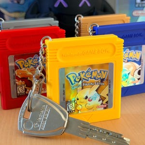 Puede incluir: Una colección de llaveros con forma de cartucho de Nintendo Game Boy en rojo, amarillo, azul y naranja, cada uno con una etiqueta de juego Pokémon. Una llave de metal plateado está unida al llavero amarillo Pikachu Edition mediante una cadena.