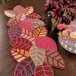 Pode incluir: Um caminho de mesa decorativo com um conjunto de formas de folhas sobrepostas. As folhas são intrincadamente cobertas com contas de vidro em tons de rosa choque, vermelho profundo, dourado metálico e preto, num padrão de folhagem natural.