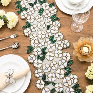 Può includere: Un runner da tavola rettangolare caratterizzato da un intricato motivo di fiori di perline bianche con centri iridescenti e foglie di perline verde scuro. Il disegno floreale è densamente disposto lungo tutta la lunghezza, creando una superficie strutturata.
