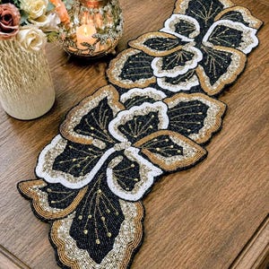 May include: A rectangular table runner featuring an intricate floral pattern crafted from small beads in black, white, and metallic gold. The design consists of repeating scalloped flower shapes with layered petals, creating a textured surface.