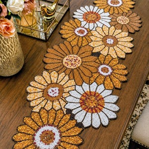 May include: A beaded sunflower table runner in shades of gold, brown, and white. The runner is made of small beads and is shaped like large flowers. The runner is on a dark wood table.