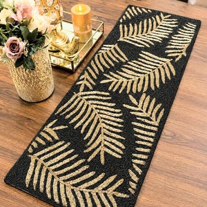 May include: A rectangular table runner featuring a black background with a repeating pattern of gold beaded fern leaves. The runner is crafted with intricate beadwork, showcasing a contrast between the dark base and the metallic gold foliage design.