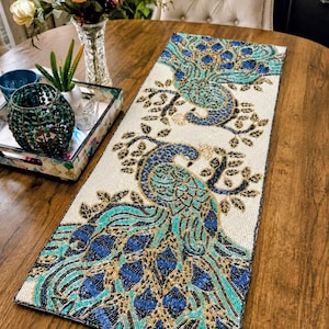 May include: A rectangular table runner featuring an intricate beaded design of two peacocks. The peacocks are rendered in shades of turquoise, royal blue, and gold beads against a cream-colored background, with detailed floral branches.