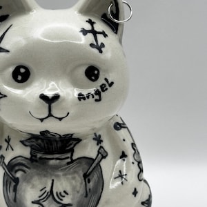 Peut inclure: Une figurine de chat en céramique blanche ornée d'illustrations à l'encre noire représentant un cœur, des croix, des étoiles et le mot angel écrit sur la joue. Le chat a une finition brillante et un petit anneau en métal argenté à l'oreille.