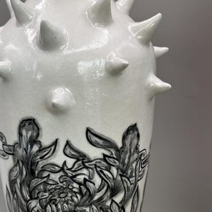 Könnte beinhalten: Eine weiße Keramikvase mit glänzender Oberfläche, die im oberen Bereich mit mehreren hervorstehenden Stacheln versehen ist und im unteren Teil ein detailliertes, handgemaltes schwarz-weißes Blumenmuster mit komplexen Blattstrukturen aufweist.