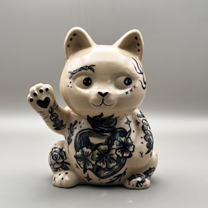 Może przedstawiać: Beżowa ceramiczna figurka kota na szczęście z czarnymi ilustracjami w stylu tatuażu przedstawiającymi kwiaty, serce, świecę i różne symbole na całym ciele. Kot siedzi z jedną podniesioną łapą, prezentując szczegółowe rysunki liniowe.