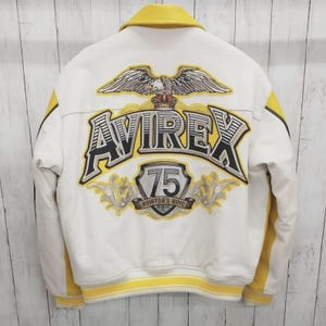 May include: White leather jacket with yellow accents and a yellow collar. The back features a large embroidered design with the word "AVIREX", an eagle, and the number "75". The jacket has a ribbed yellow hem and cuffs.