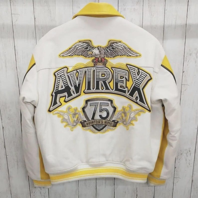 May include: White leather jacket with yellow accents and a yellow collar. The back features a large embroidered design with the word "AVIREX", an eagle, and the number "75". The jacket has a ribbed yellow hem and cuffs.