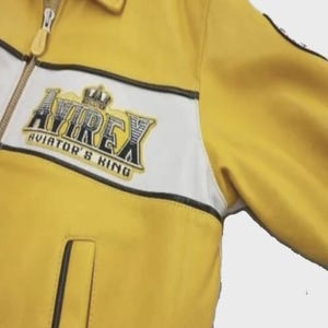 May include: Yellow jacket with white and black accents. The jacket features a zipper closure and a patch on the chest with the text "AVIREX AVIATOR'S KING" in black and gold. The jacket has a classic design.