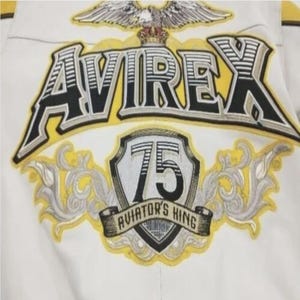 May include: Close-up of a white jacket with embroidered details. The jacket features the word "AVIREX" in a bold, outlined font, with a yellow and black color scheme. Below, a shield displays "75" and "AVIATOR'S KING". An eagle with a crown is at the top.