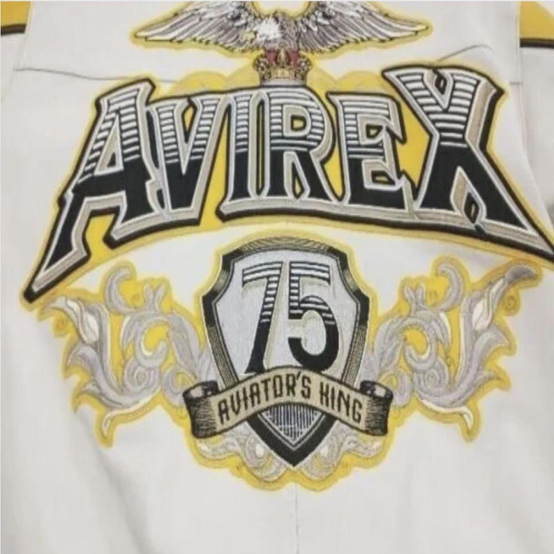 May include: Close-up of a white jacket with embroidered details. The jacket features the word "AVIREX" in a bold, outlined font, with a yellow and black color scheme. Below, a shield displays "75" and "AVIATOR'S KING". An eagle with a crown is at the top.