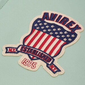 K&ouml;nnte beinhalten: Gestickter Aufn&auml;her mit einem Schilddesign, roten und wei&szlig;en Streifen und einem blauen Sternenfeld. Der Aufn&auml;her enth&auml;lt den Text "AVIREX", "ESTABLISHED 1975" und "N.Y." in roter und blauer Stickerei.