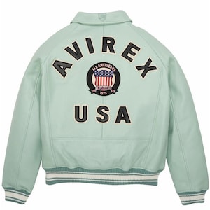 K&ouml;nnte beinhalten: Hellgr&uuml;ne Lederjacke mit dem Wort "AVIREX" in schwarzen Blockbuchstaben &uuml;ber den Schultern. Darunter befindet sich ein kreisf&ouml;rmiger Aufn&auml;her mit den Worten "ALL AMERICANS ESTABLISHED 1975" und einem US-Flaggen-Design. Das Wort "USA" steht darunter.