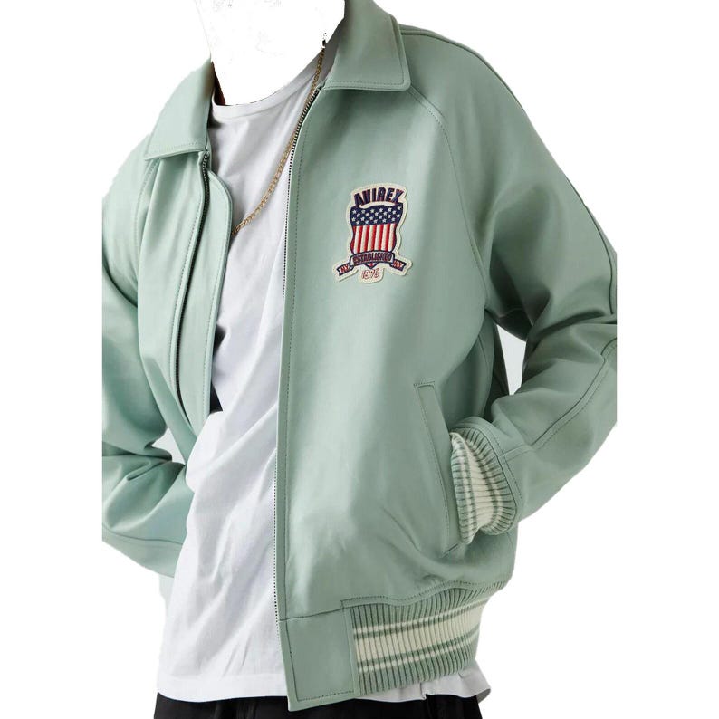 K&ouml;nnte beinhalten: Hellgr&uuml;ne Bomberjacke mit Rei&szlig;verschluss und Kragen. Die Jacke hat einen Aufn&auml;her mit dem Text "AVIREX" und einem amerikanischen Flaggen-Design. Die Manschetten und der Saum haben ein Streifenmuster.