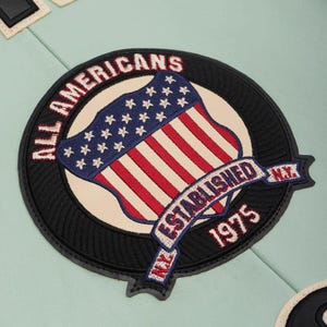 K&ouml;nnte beinhalten: Runder, gestickter Aufn&auml;her mit einem Schilddesign der amerikanischen Flagge, mit dem Text "ALL AMERICANS" und "ESTABLISHED 1975 N.Y." in Rot und Wei&szlig;. Der Aufn&auml;her hat einen schwarzen Rand und liegt auf einem hellblauen Hintergrund.
