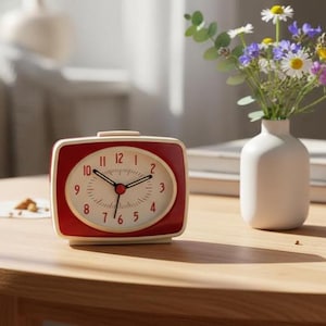 Könnte beinhalten: Ein roter und cremefarbener Retro-Wecker mit weißem Zifferblatt und schwarzen Zeigern steht auf einem Holztisch. Die Uhr hat eine quadratische Form mit abgerundeten Ecken. Eine kleine weiße Vase mit bunten Blumen steht neben der Uhr.