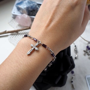 May include: A silver cross charm bracelet with purple glass beads. The bracelet is on a person's wrist.