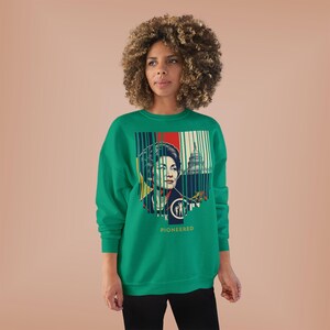 Puede incluir: Una sudadera verde con un estampado gráfico. El diseño incluye un retrato de mujer, el Capitolio de los Estados Unidos y la palabra "PIONEERED". El gráfico incorpora elementos rojos, blancos y azules. La sudadera es de manga larga y cuello redondo.
